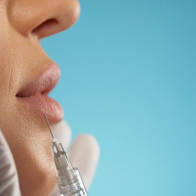 Close up of cosmetic botox injection in female lips. Side view of cosmetologist using small syringe with special liquid to make lips bigger, isolated on blue. Concept of cosmetology, beauty.